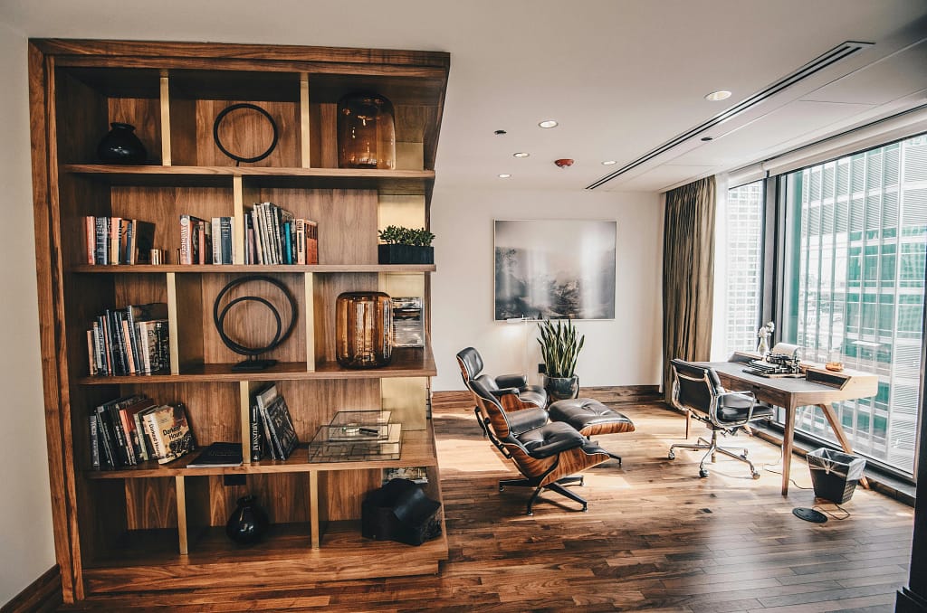 A modern office with wooden flooring, a large window, a bookshelf filled with books and decor, an ergonomic chair, and a desk with a computer.