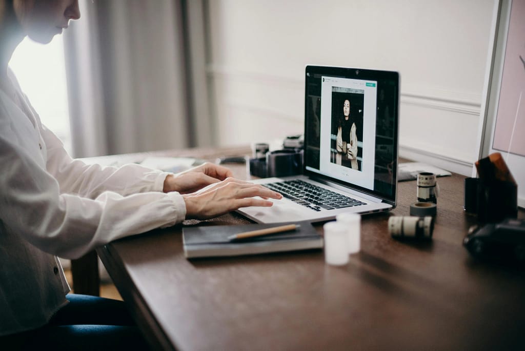 Person sitting at a desk using a laptop with photo editing software and various photography equipment placed on the table.