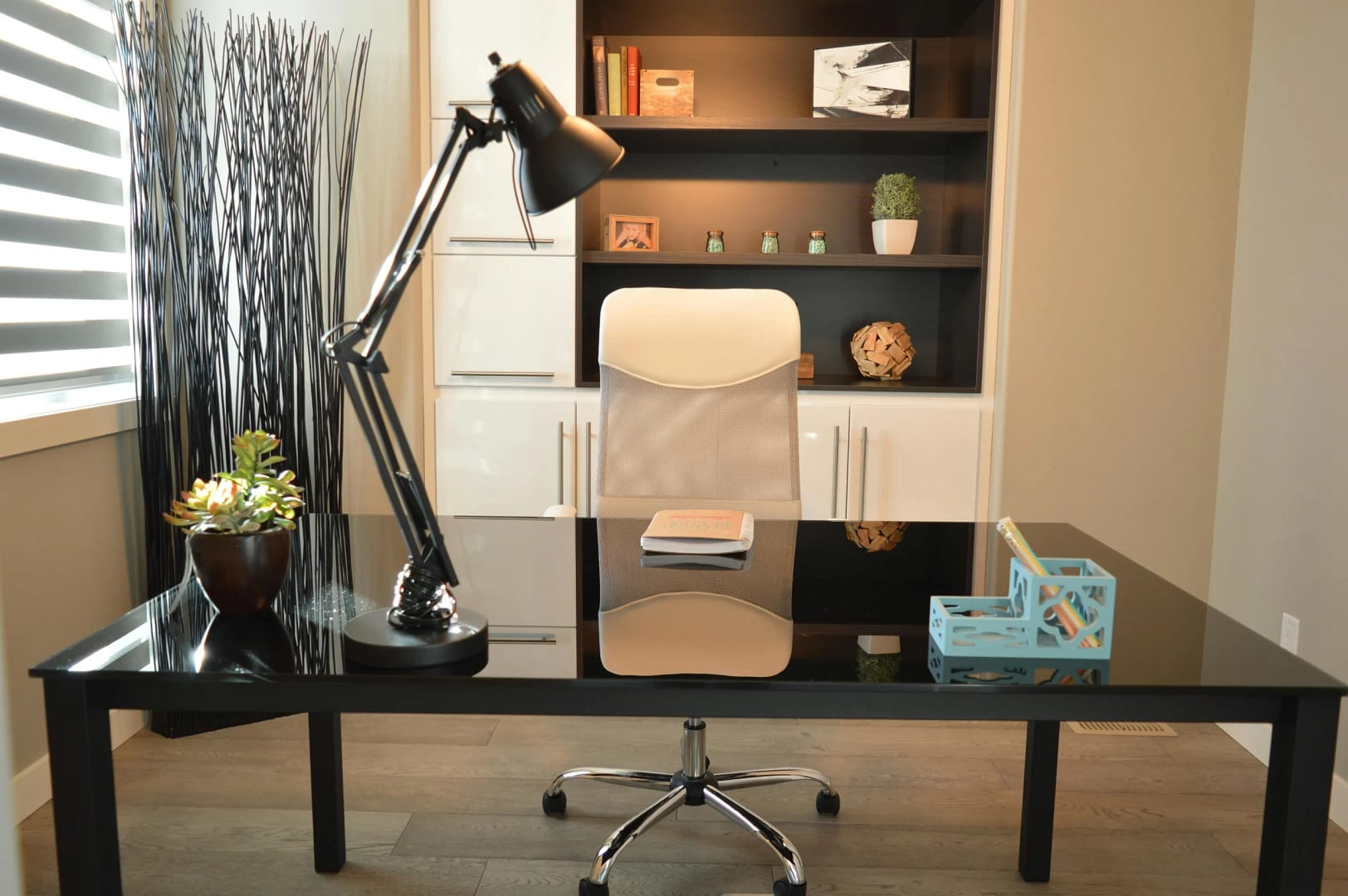 A modern home office with a black desk, white chair, desk lamp, plant, and bookshelf.