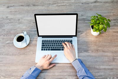 Hands typing on a laptop keyboard on a wooden desk, with a cup of coffee and a potted plant nearby.