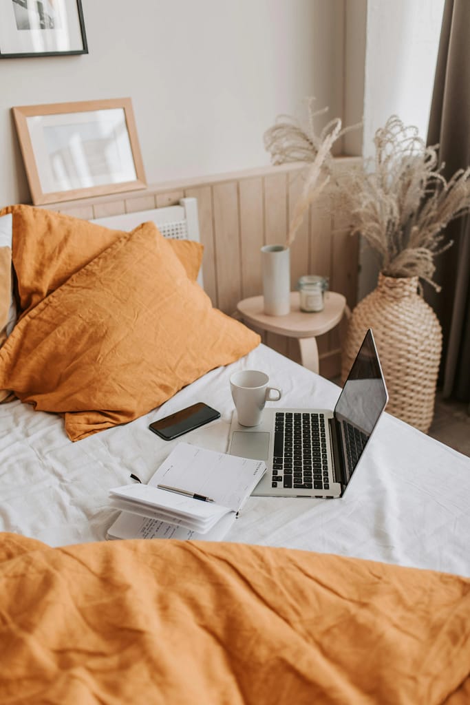 Cozy bedroom with a bed used as a workspace; featuring a laptop, documents, phone, and a cup of coffee, all on mustard-yellow bedding.