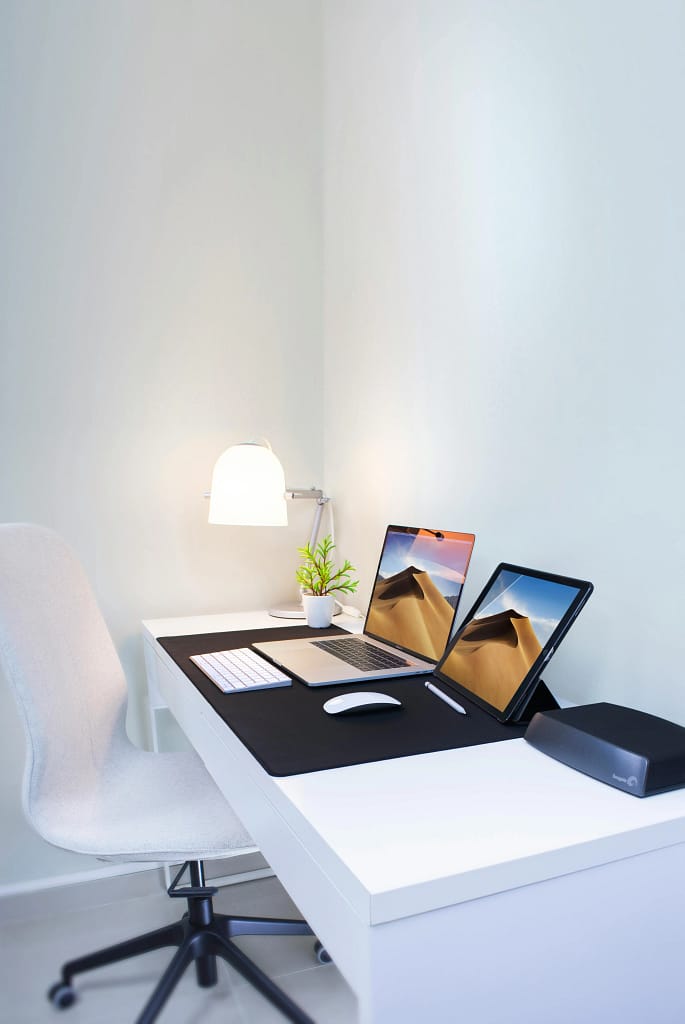 A clean and minimalistic workspace featuring a white desk, a laptop, a second computer screen, a potted plant, a desk lamp, and a white office chair.